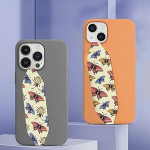May include: Two smartphone cases, one in matte gray and one in matte orange, each featuring a vertical, leaf-shaped insert on the back. The inserts are cream-colored and decorated with a repeating pattern of colorful butterflies and beetles.