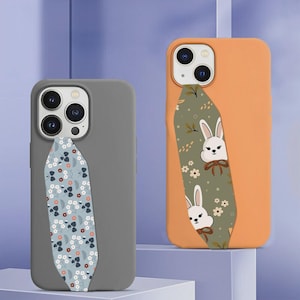 Op de afbeelding: Twee smartphonehoesjes, een matgrijs en een mat-oranje, elk voorzien van een verticale stoffen band. Het grijze hoesje heeft een blauwe band met een wit en rood bloemenpatroon, het oranje hoesje een groene band met witte konijnenillustraties.