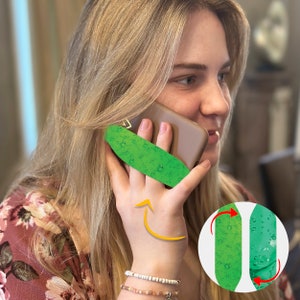 May include: A green phone grip with a textured surface that looks like a cucumber. The grip is attached to a brown phone case. The phone is being held by a person's hand.