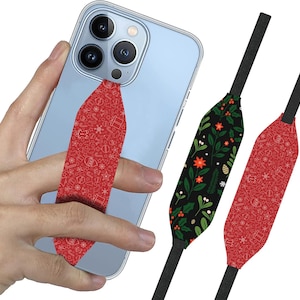 May include: A red and white patterned phone grip with a Christmas theme. The grip is attached to a light blue iPhone with a black and white Christmas floral patterned phone strap.