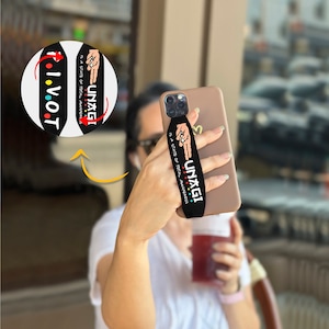 Pode incluir: Pega de telefone preta com o texto "UNAGI é um estado de consciência total" em cores do arco-íris. A pega do telefone está presa a uma capa de telefone castanho.