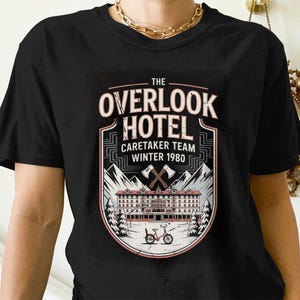 Könnte beinhalten: Schwarzes T-Shirt mit einem Grafikdesign des Overlook Hotels, mit dem Text "The Overlook Hotel Caretaker Team Winter 1980". Das Design zeigt gekreuzte Äxte, Berge und ein Fahrrad.