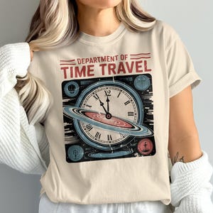 May include: Cream-colored t-shirt featuring a vintage-style graphic. The design includes a clock with a planet-like ring, and the words "DEPARTMENT OF TIME TRAVEL" in red. The clock hands point to nearly 12 o'clock.