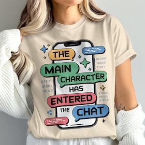 May include: Beige t-shirt with a smartphone graphic and the text "THE MAIN CHARACTER HAS ENTERED THE CHAT." The design incorporates speech bubbles, stars, and binary code. A casual wear item.