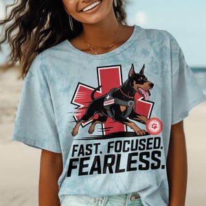 May include: A light blue t-shirt featuring a red medical symbol with a black and brown dog running through it. The text "FAST. FOCUSED. FEARLESS." is printed below the symbol.