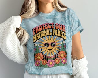 Puede incluir: Una camiseta azul claro tie-dye con un diseño gráfico retro que presenta un sol sonriente con gafas de sol y el texto "Protect Our National Parks".