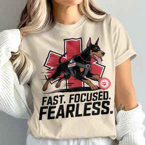 May include: Beige t-shirt featuring a Doberman Pinscher graphic in action, wearing a service vest, set against a red cross. The text "FAST. FOCUSED. FEARLESS." is printed below the dog. A paw print emblem is on the right.