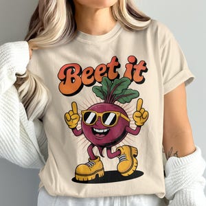 Peut inclure: T-shirt beige avec un personnage de betterave de dessin animé rétro portant des lunettes de soleil et des chaussures jaunes, avec le texte "Beet it" en orange et marron. La betterave fait un signe de la main.