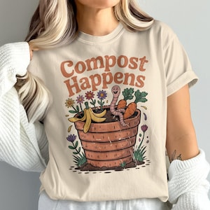 May include: Cream-coloured t-shirt featuring the words "Compost Happens" in orange. The illustration depicts a smiling worm in a plant pot with carrots, flowers, and a banana peel. The design has a retro cartoon style.