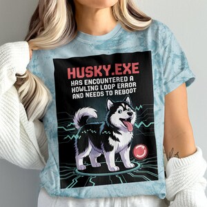 May include: A light blue t-shirt with a black and white illustration of a husky dog. The text on the shirt reads "HUSKY.EXE HAS ENCOUNTERED A HOWLING LOOP ERROR AND NEEDS TO REBOOT".