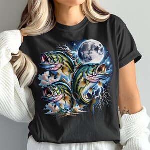 Op de afbeelding: Zwart T-shirt met een kleurrijke afbeelding van drie baarsvissen met open mond, tegen een achtergrond van een volle maan, bliksem en wolken. Het ontwerp gebruikt tinten blauw, groen en geel.