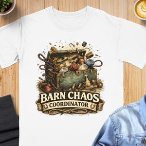 May include: White t-shirt featuring a detailed illustration of an open trunk overflowing with equestrian equipment and supplies. The text "BARN CHAOS COORDINATOR" is displayed on a banner below the trunk. The design includes various colors and textures.