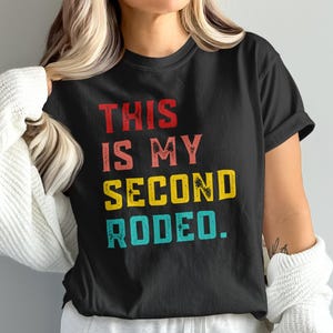 Könnte beinhalten: Schwarzes T-Shirt mit dem Text "THIS IS MY SECOND RODEO" in Rot, Rosa, Gelb und Türkis. Der Text hat einen Used-Look im Vintage-Stil. Das Shirt ist ein Freizeitkleidungsstück.