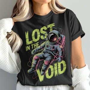 May include: Black t-shirt featuring a colorful illustration of an astronaut floating in space. The text "LOST IN THE VOID" is in a distressed, neon green font. The astronaut is wearing a dark blue spacesuit with pink and yellow accents.