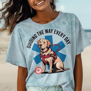 Könnte beinhalten: Ein hellblaues T-Shirt mit einem Aufdruck eines gelben Labrador Retrievers, der ein rotes Assistenzhund-Geschirr trägt. Der Hund steht vor einem blauen Stern-des-Lebens-Symbol. Der Text "Guiding the way every day" ist auf das Shirt gedruckt.