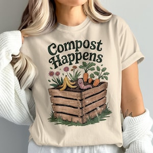 May include: Beige t-shirt with the text "Compost Happens" in green. The graphic shows a wooden compost bin with a smiling worm, carrots, flowers, and a banana peel. The shirt has short sleeves.
