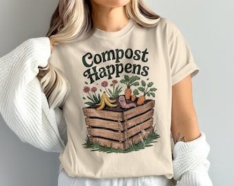 May include: Beige t-shirt with the text "Compost Happens" in green. The graphic shows a wooden compost bin with a smiling worm, carrots, flowers, and a banana peel. The shirt has short sleeves.