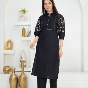 May include: A black knee-length tunic with a mandarin collar, button-down front placket, and three-quarter length sleeves featuring white floral embroidery. The garment is paired with matching black trousers. The text HodgePodgeYarn is visible.