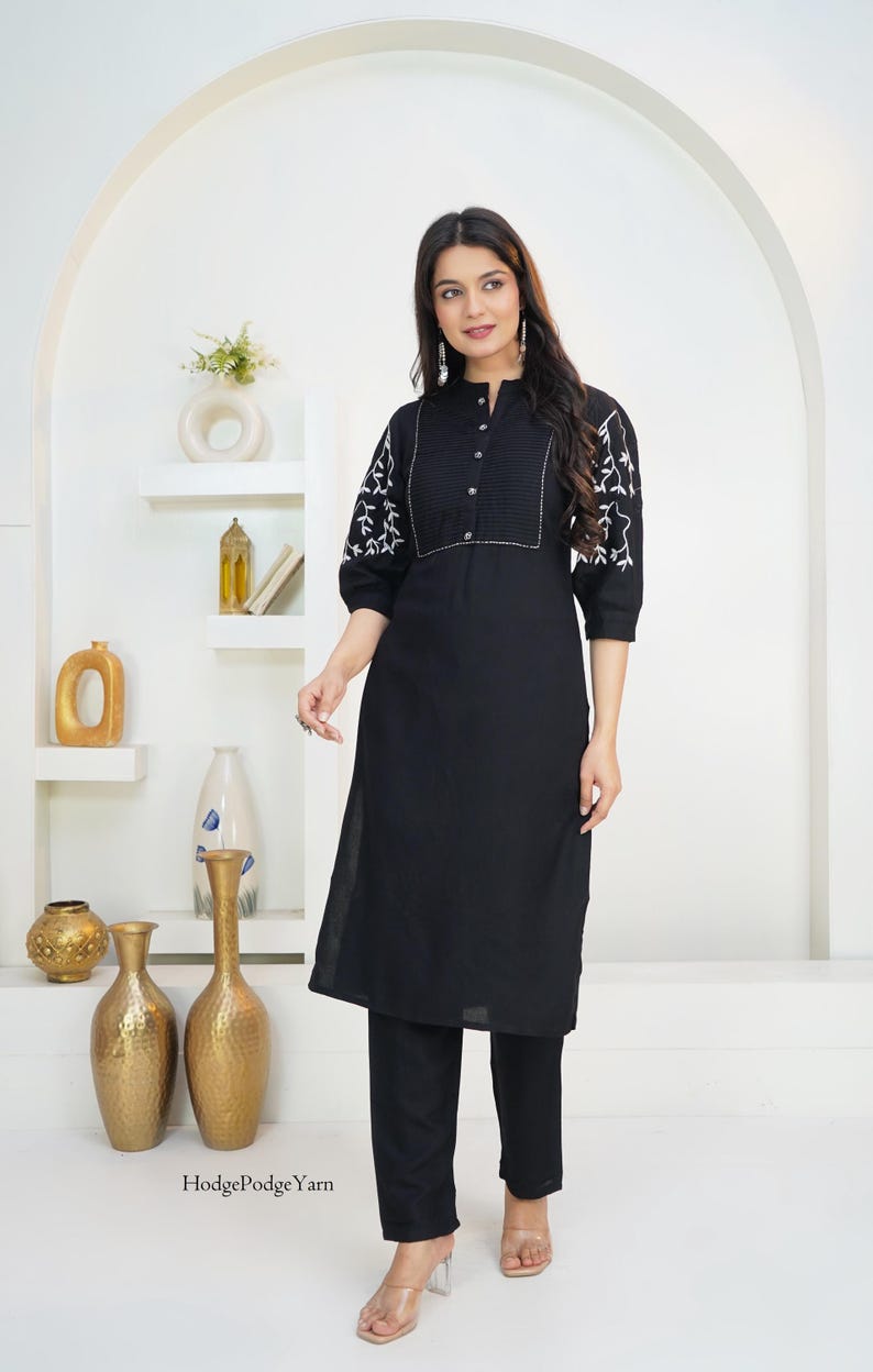 May include: A black knee-length tunic with a mandarin collar, button-down front placket, and three-quarter length sleeves featuring white floral embroidery. The garment is paired with matching black trousers. The text HodgePodgeYarn is visible.