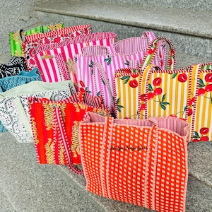 May include: A selection of tote bags in a variety of patterns, including stripes, floral, and gingham. The bags feature bright colours such as pink, red, yellow, and green. Some bags have cherry and tiger prints. The bags are displayed on concrete steps.