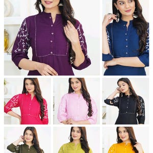 May include: A collection of solid-colored tunics featuring a mandarin collar, a button-down front placket with vertical pleats, and three-quarter length sleeves adorned with white floral embroidery. Available in purple, blue, pink, black, olive, and yellow.