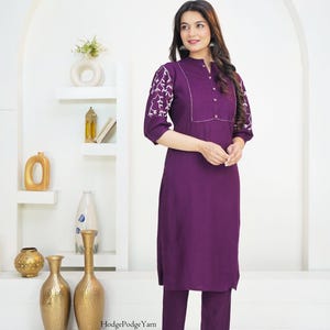 May include: A deep purple tunic and matching trousers set. The tunic features three-quarter length sleeves with white floral embroidery, a pleated chest panel, and a button-down collar. The text HodgePodgeYarn is visible near the bottom hem.