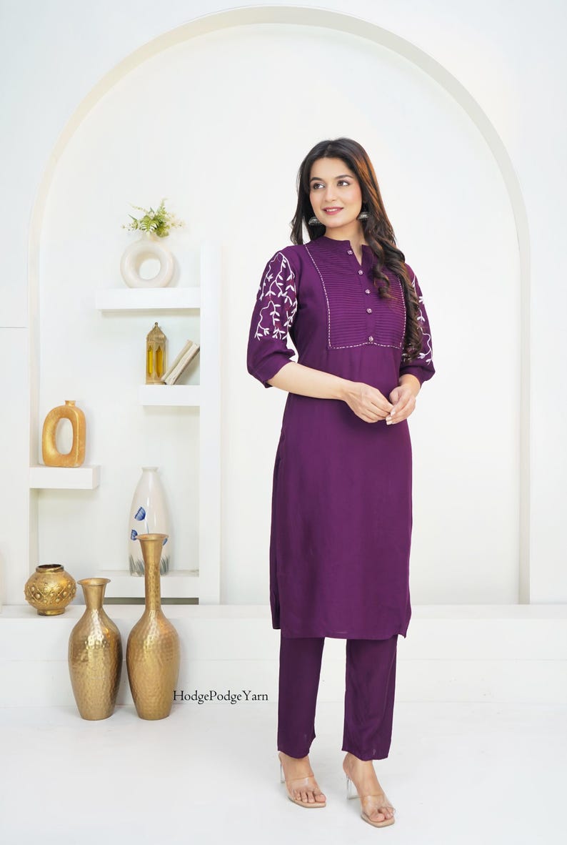 May include: A deep purple tunic and matching trousers set. The tunic features three-quarter length sleeves with white floral embroidery, a pleated chest panel, and a button-down collar. The text HodgePodgeYarn is visible near the bottom hem.