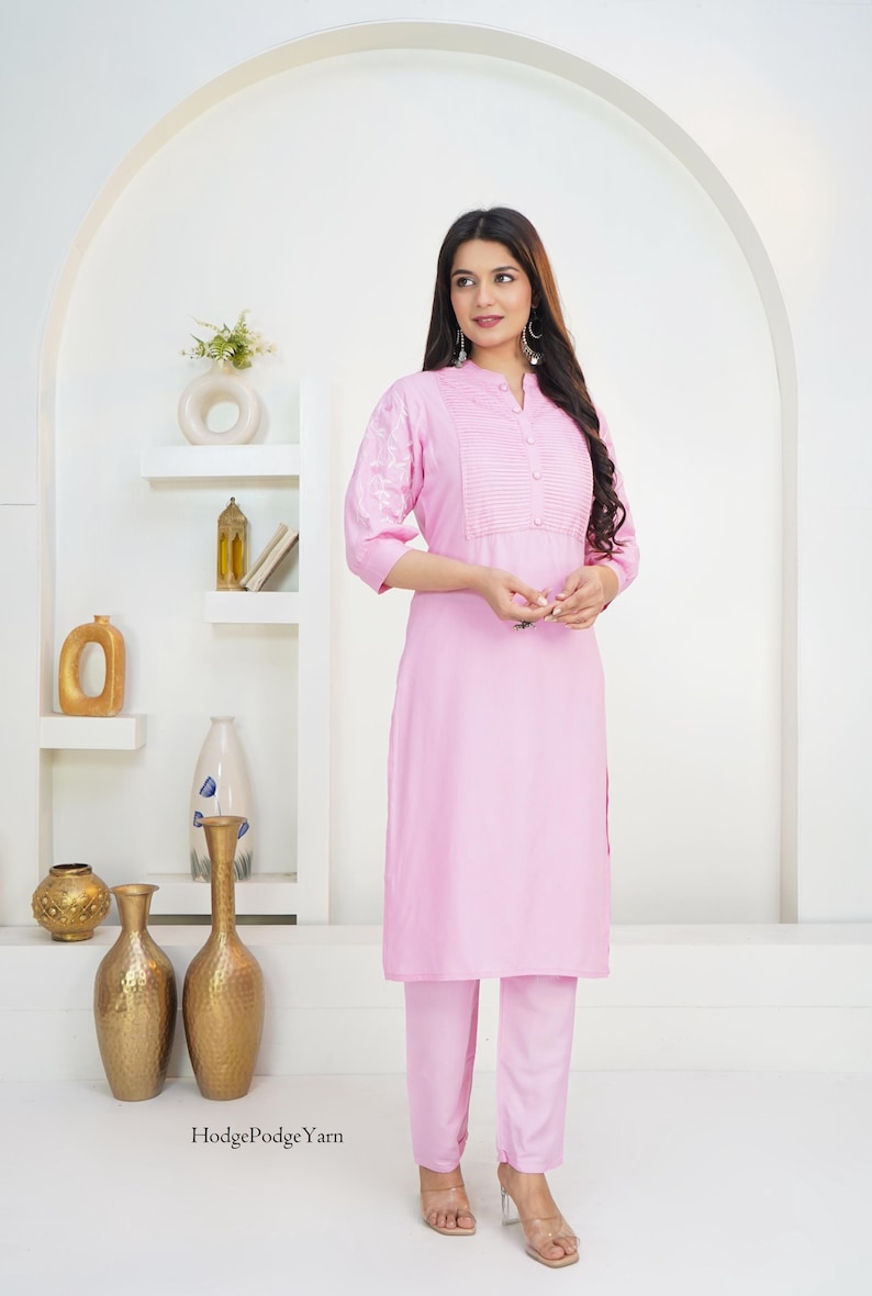 May include: A light pink long-sleeved tunic with a mandarin collar, a button-down placket with vertical pleats, and white floral embroidery on the sleeves, paired with matching light pink straight-leg trousers. The text HodgePodgeYarn is visible.