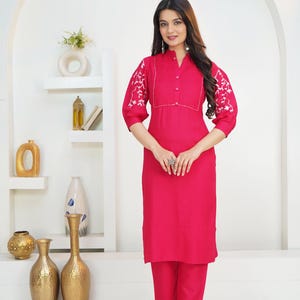 May include: A vibrant fuchsia kurta set featuring a straight-cut tunic with a mandarin collar, button-down placket, and pleated chest detail. The three-quarter length sleeves are adorned with white floral embroidery. Matching fuchsia trousers.