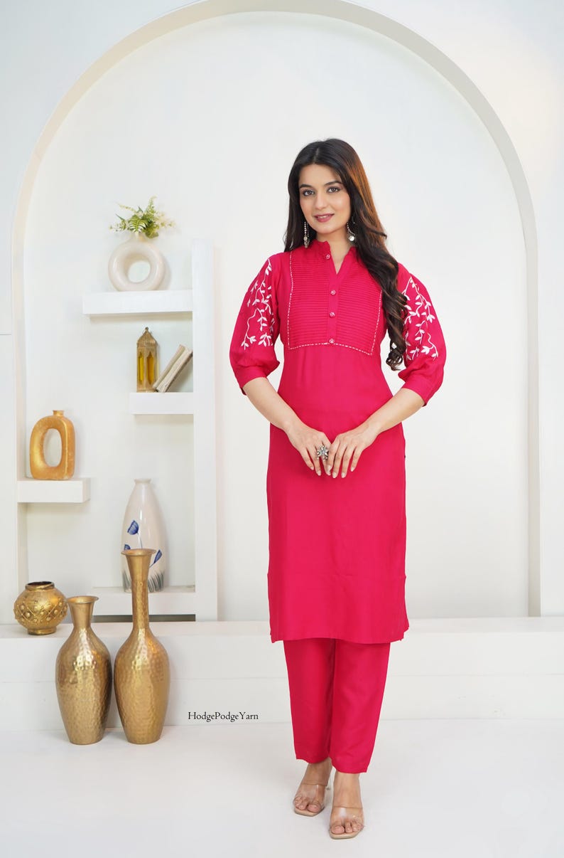 May include: A vibrant fuchsia kurta set featuring a straight-cut tunic with a mandarin collar, button-down placket, and pleated chest detail. The three-quarter length sleeves are adorned with white floral embroidery. Matching fuchsia trousers.