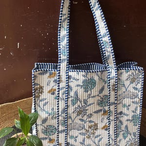 以下が含まれることがあります： 白地に青、緑、黄色の花柄が施されたキルティングトートバッグ。バッグには青いストライプのトリムとハンドルが付いています。「Hodge Podge Yarn」の文字が上部に表示されています。買い物に最適です。