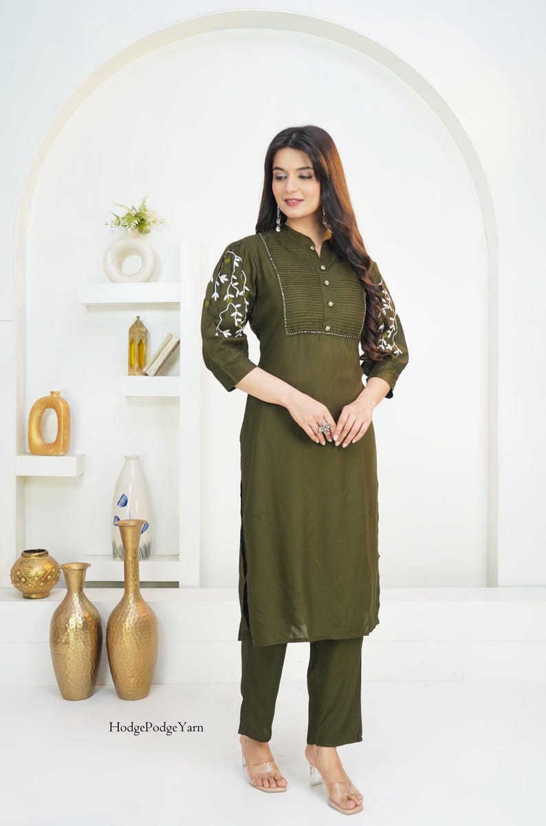 May include: An olive green kurta set featuring a long-sleeved tunic with a pleated bodice, button-down front, and white floral embroidery on the sleeves, paired with matching straight-leg trousers. The text HodgePodgeYarn is visible at the bottom.