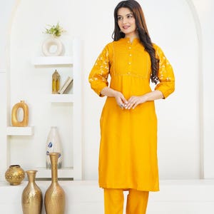 May include: A mustard yellow long-sleeved tunic and matching trousers set. The tunic features a mandarin collar, a central button placket with vertical pleats, and white floral embroidery on the sleeves. The text HodgePodgeYarn is visible.