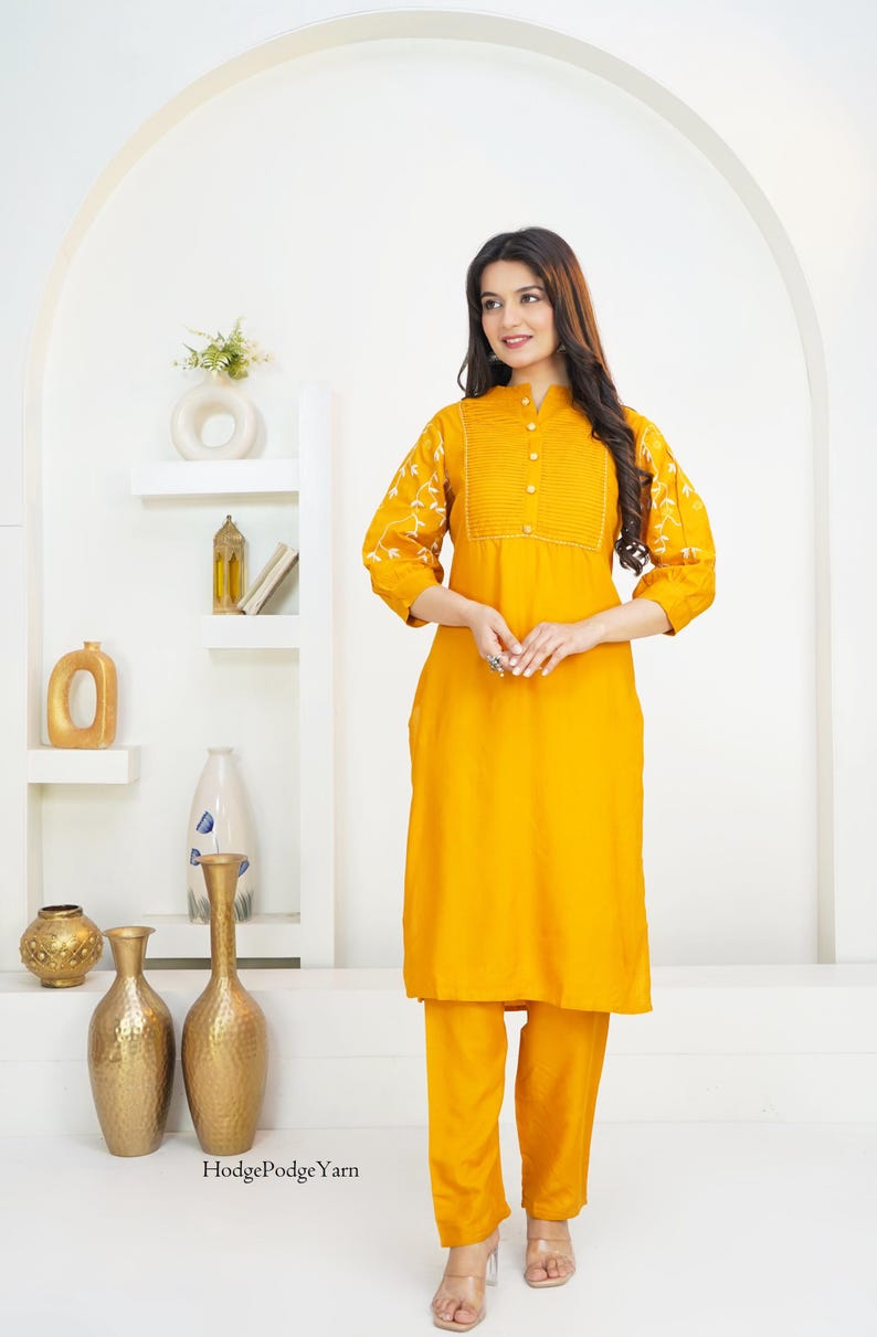 May include: A mustard yellow long-sleeved tunic and matching trousers set. The tunic features a mandarin collar, a central button placket with vertical pleats, and white floral embroidery on the sleeves. The text HodgePodgeYarn is visible.