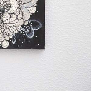 Pode incluir: Uma pintura em tela preta com desenhos abstratos brancos e cinzentos. A pintura &eacute; emoldurada e pendurada em uma parede branca.