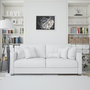 Pode incluir: Um sof&aacute; branco com duas almofadas fica em uma sala branca com prateleiras brancas e uma obra de arte emoldurada em preto e branco na parede. As prateleiras est&atilde;o cheias de livros e itens decorativos.