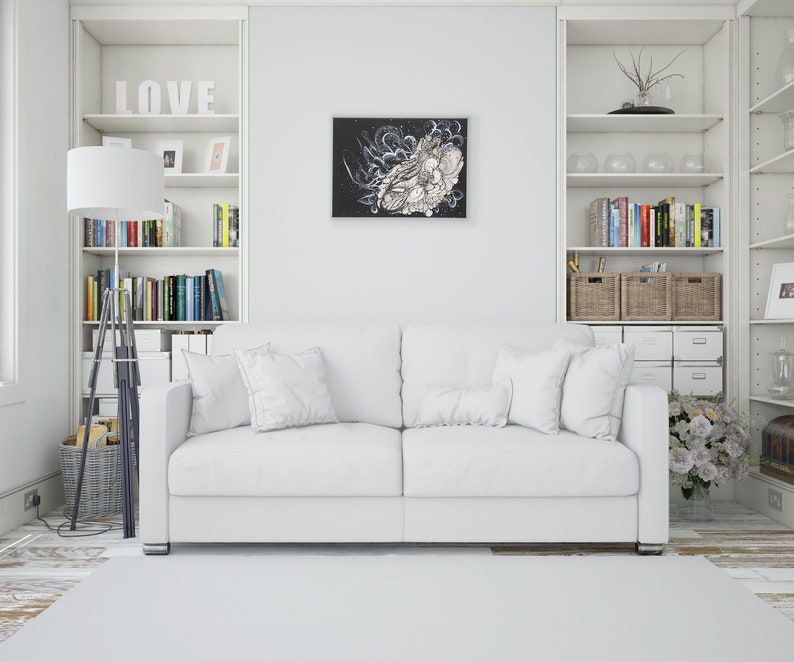 Pode incluir: Um sof&aacute; branco com duas almofadas fica em uma sala branca com prateleiras brancas e uma obra de arte emoldurada em preto e branco na parede. As prateleiras est&atilde;o cheias de livros e itens decorativos.