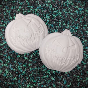 May include: Two white ceramic pumpkin-shaped decorations with carved jack-o'-lantern faces. The pumpkins are on a black and green background.