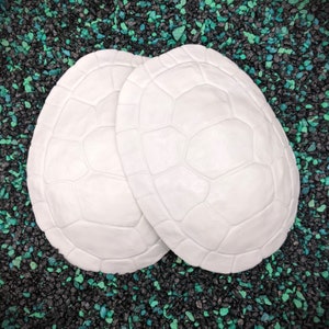 May include: Two white turtle shell moulds, with a textured surface, on a black and green background.