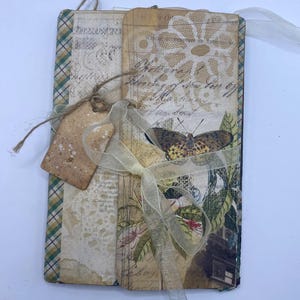 May include: A handmade junk journal featuring a vintage-style cover with a butterfly illustration, botanical prints, and cursive script text. The journal is bound with a sheer cream ribbon and includes a small, speckled brown paper tag tied with twine.