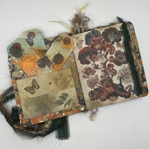 May include: Open journal with botanical illustrations of mushrooms and other plants. The journal has a decorative cover with tags and a pocket. The tags have orange and blue splattered designs. A butterfly and leaves are printed on the pocket.