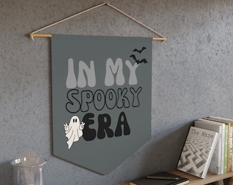 Può includere: Uno stendardo grigio con il testo "In My Spooky Era" in un font retrò. Un fantasma bianco con le braccia alzate è a sinistra del testo. Due pipistrelli neri volano sopra il testo. Lo stendardo è appeso a un bastone di legno.