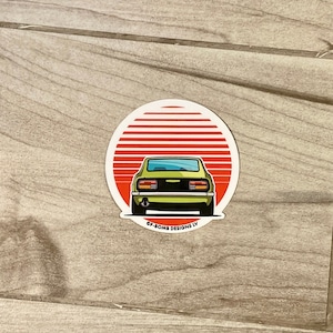 May include: A yellow car sticker with a red and white sunset behind it. The car is a vintage Datsun 240Z. The sticker says "SF BOMB DESIGNS IV" at the bottom.