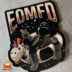 Peut inclure: Un autocollant noir et blanc avec un astronaute de dessin animé chevauchant une licorne dans l'espace. L'astronaute tient une boisson et l'autocollant dit "COMFD".