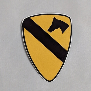 May include: A yellow and black military patch with a horse head silhouette. The patch is shaped like a shield with a black diagonal stripe.