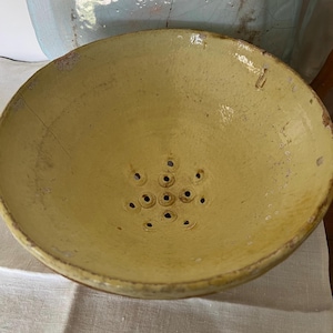 May include: A round, yellow glazed ceramic colander featuring a wide, open bowl shape with a cluster of drainage holes at the center. The surface shows visible signs of wear, including fine cracks in the glaze and small chips along the rim.