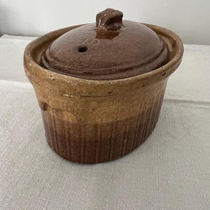 May include: An antique brown ceramic casserole dish with a lid. The dish has a ribbed exterior and a handle on the lid. The color of the dish transitions from a light tan to a darker brown. A small hole is visible on the lid.