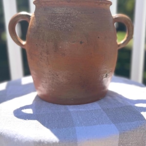 May include: A rustic, terracotta-colored ceramic pot with two loop handles. The pot has a wide, rounded body and a slightly flared rim. It sits on a checkered tablecloth, with a bright outdoor background.
