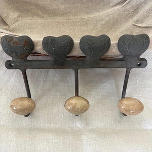 May include: A wall-mounted coat rack made of dark, weathered metal featuring four heart-shaped decorative elements along the top bar. Three downward-curving hooks extend from the bar, each finished with a smooth, round, beige-toned ceramic knob.