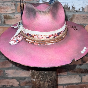 May include: A pink felt cowboy hat with a floral patterned band, a feather, and a denim ribbon. The hat has a distressed look with white paint splatters.